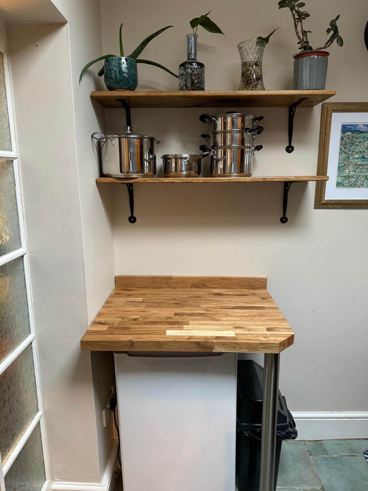 Kitchen corner with wooden shelves holding pots, pans, and plants, above a small fridge and a wooden countertop.