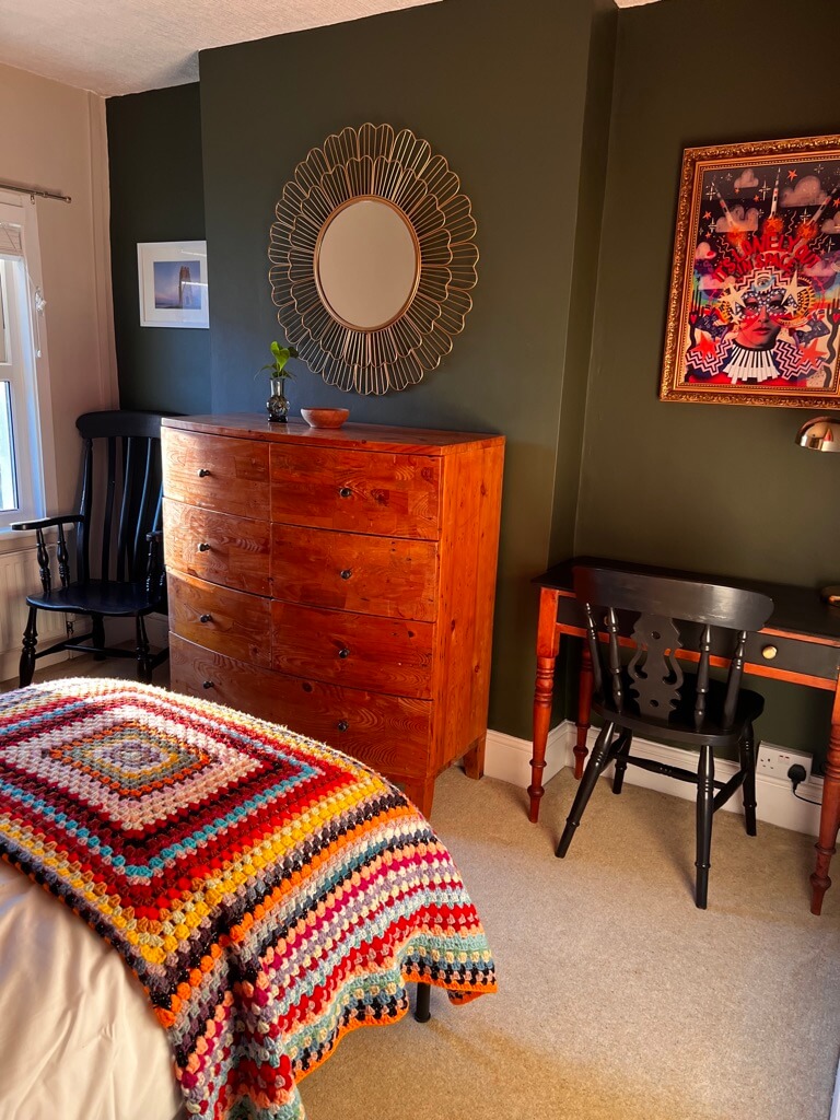 Cozy bedroom with a colorful crochet blanket on the bed, a wooden dresser, a wall mirror, a small desk with a chair, and vibrant artwork on dark green walls.