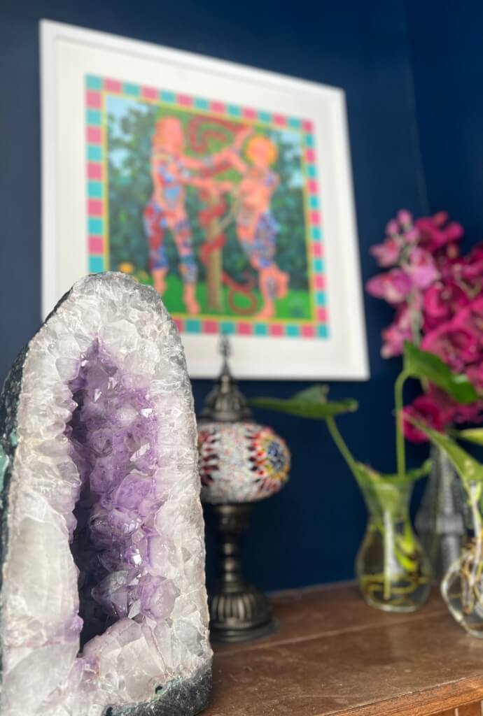 Amethyst geode on a wooden table with vases of pink flowers, a decorative lamp, and a framed painting on a blue wall.