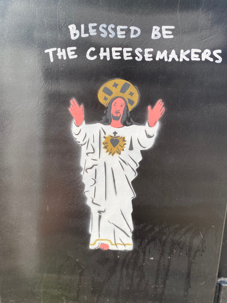 Illustration of a religious figure in white robes with a halo, arms raised. The text above reads "Blessed Be the Cheesemakers."