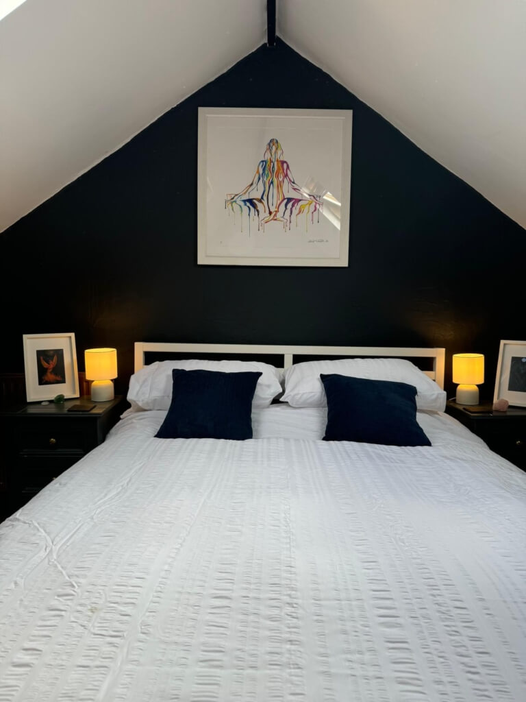 Cozy attic bedroom with a white bed, blue accent pillows, and lit lamps on dark bedside tables. A colorful art piece hangs on the slanted, dark wall above the bed.