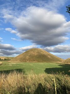 Silbury Hill under a blue sky with large, fluffy clouds, surrounded by fields and vegetation.