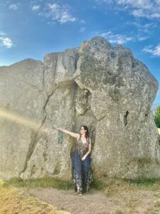 Louise Carron Harris in a patterned dress stands beside a large, weathered stone under a blue sky with scattered clouds, bathed in sunlight.