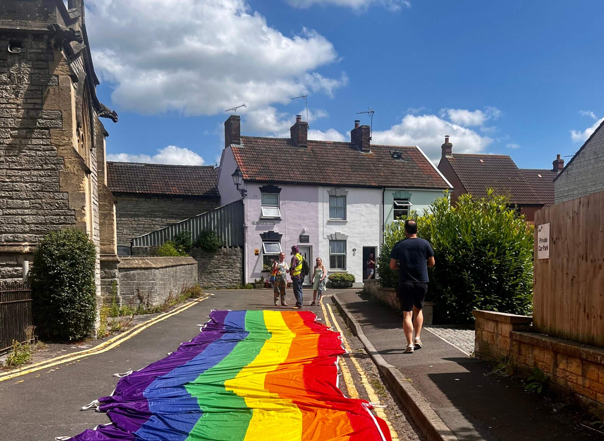 A large rainbow flag is spread across a narrow street in a quaint village with a white house in the background, under a partly cloudy sky.