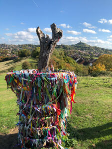 A tree stump wrapped in multicolored ribbons stands in a grassy landscape with a hill and town in the background under a blue sky with scattered clouds.