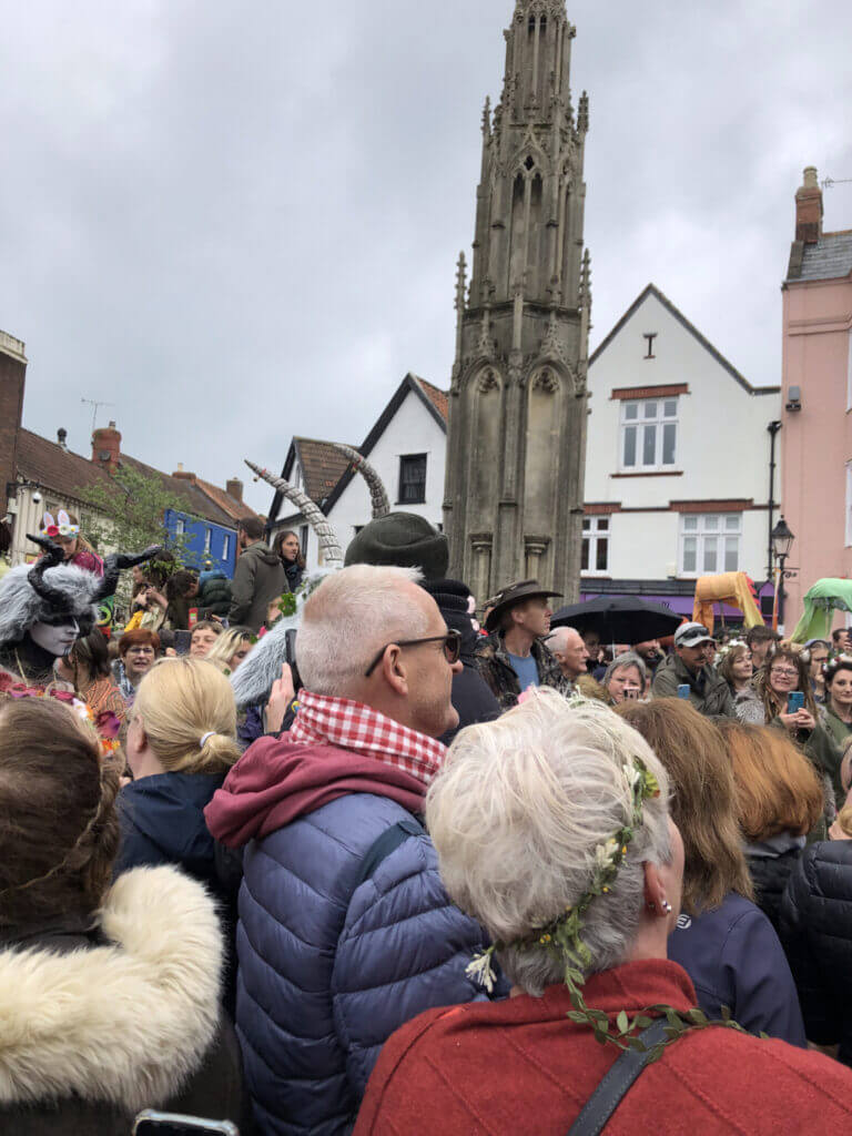 A crowd gathers in a town square, surrounded by historic buildings and a tall stone monument. People wear festive clothing, including hats and wreaths. The sky is overcast, creating a lively yet subdued atmosphere.
