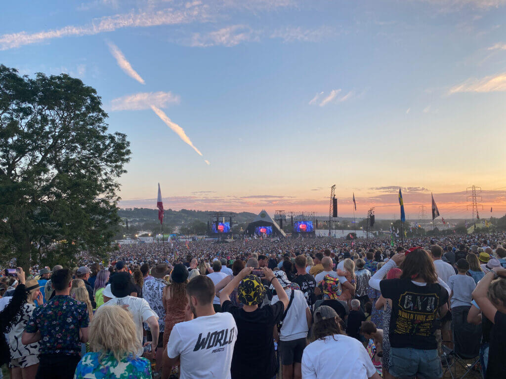 A large outdoor concert at sunset with a crowd of people facing the stage, framed by trees and flags under a colorful sky.