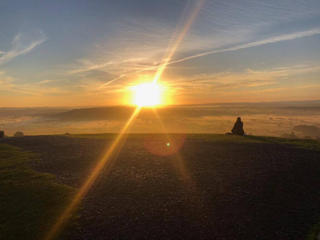 Sunrise over a misty landscape, with a bright sun casting rays across the sky and a lone rock visible on the ground.