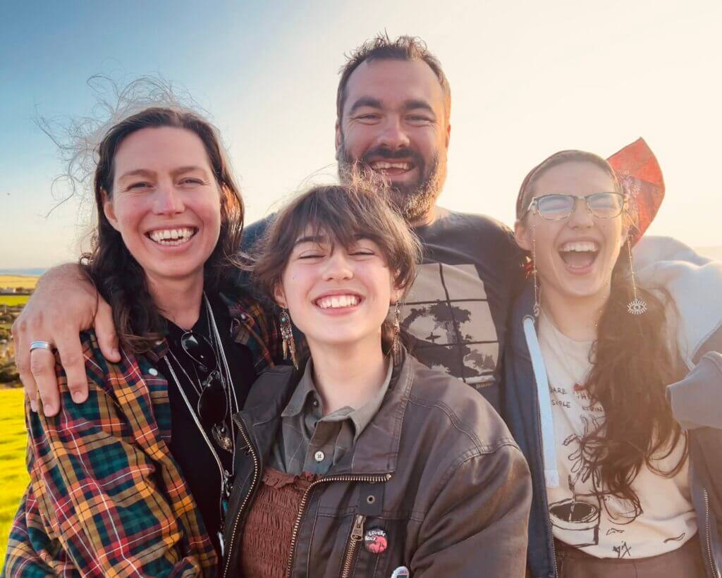 A joyful group of four people smiling broadly outdoors, with sunlight illuminating their faces against a clear sky.