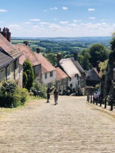 Cobblestone street in Shaftesbury descending with quaint cottages on either side, lush greenery, and a clear sky in the background.