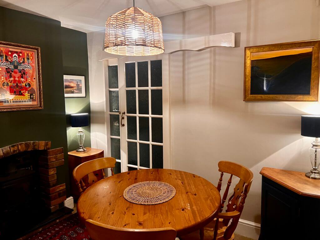 A cozy dining room with a round wooden table and chairs, illuminated by a woven light fixture. The walls feature two framed artworks, and a door with glass panes leads to another room. Warm lighting enhances the inviting atmosphere.