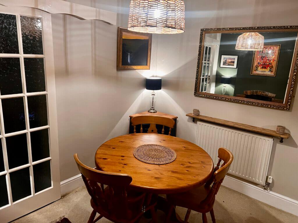 Cozy dining area with a round wooden table and four chairs, set under a woven pendant light. A large mirror and a small lamp add warmth to the inviting space.