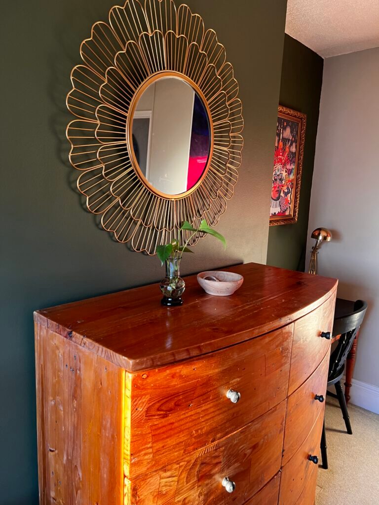 Wooden dresser with four drawers, topped with a small vase and bowl, sits against a dark wall. Above it is a decorative round mirror with a sunburst frame. A chair and artwork are partially visible in the background.
