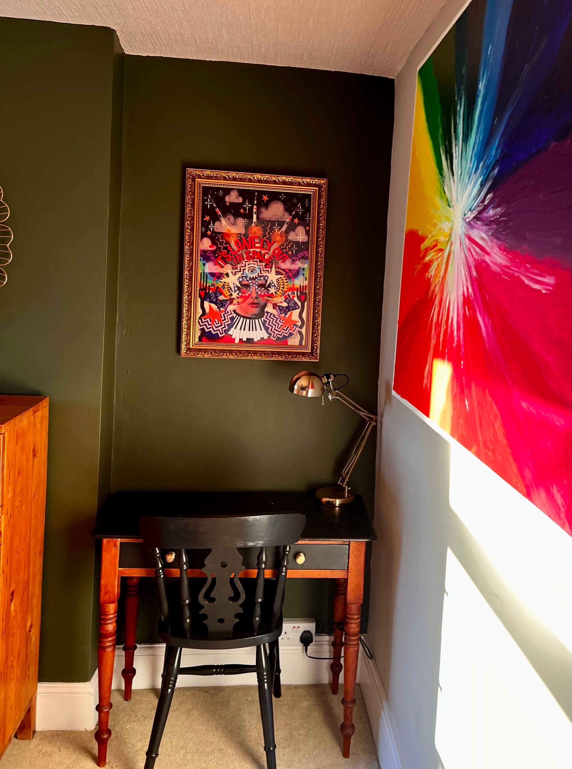 A small home office nook with a dark wooden desk and chair, lit by a desk lamp. The wall is decorated with a colorful abstract painting and a framed picture of dogs playing cards. Bright natural light illuminates the room.
