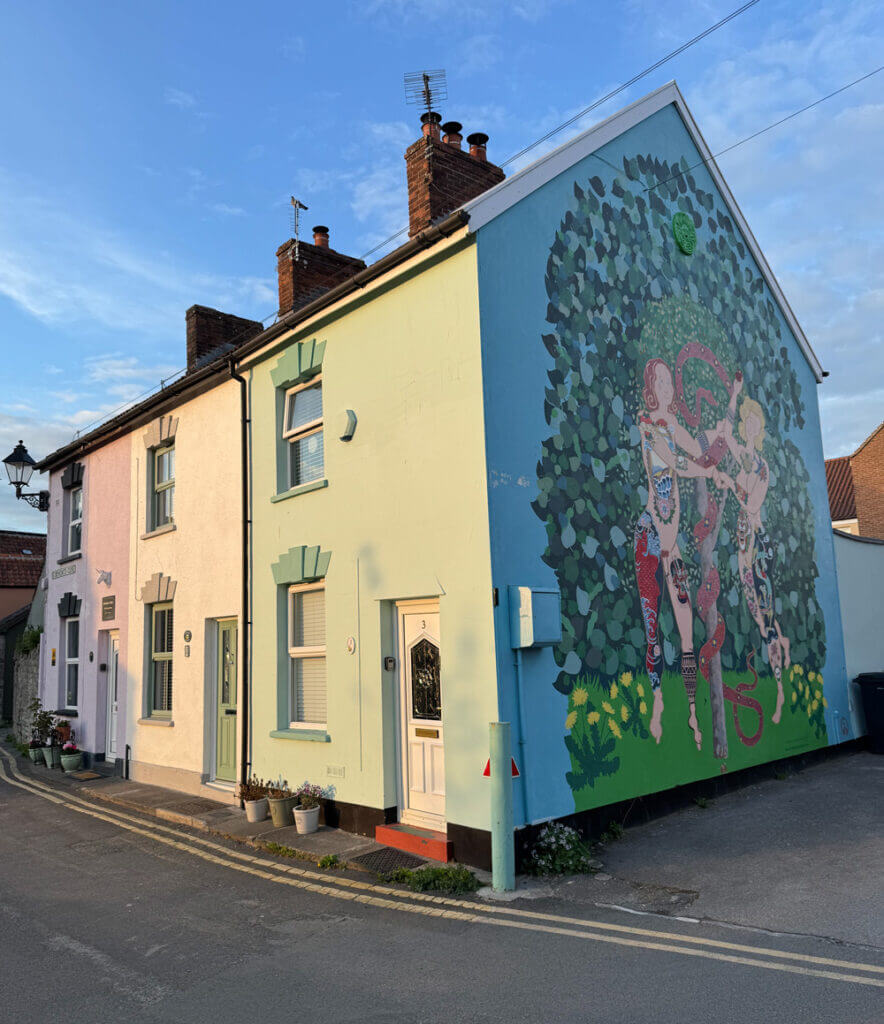 Colorful houses with a large wall mural depicting two people among foliage, situated on a sunny street corner.