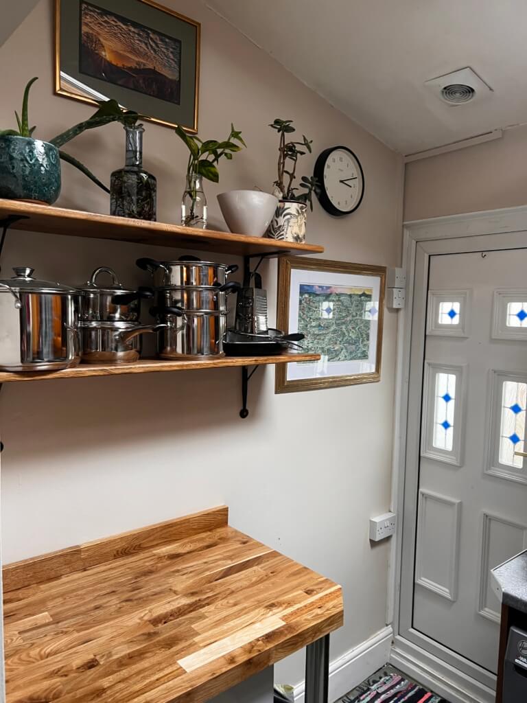 A small kitchen corner with a wooden countertop and wall shelves holding plants, pots, and jars. A clock and framed pictures are on the wall, and a white door with decorative glass panels is visible.