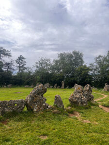 Ancient standing stones arranged in a circle on a grassy field under a cloudy sky, with trees in the background.