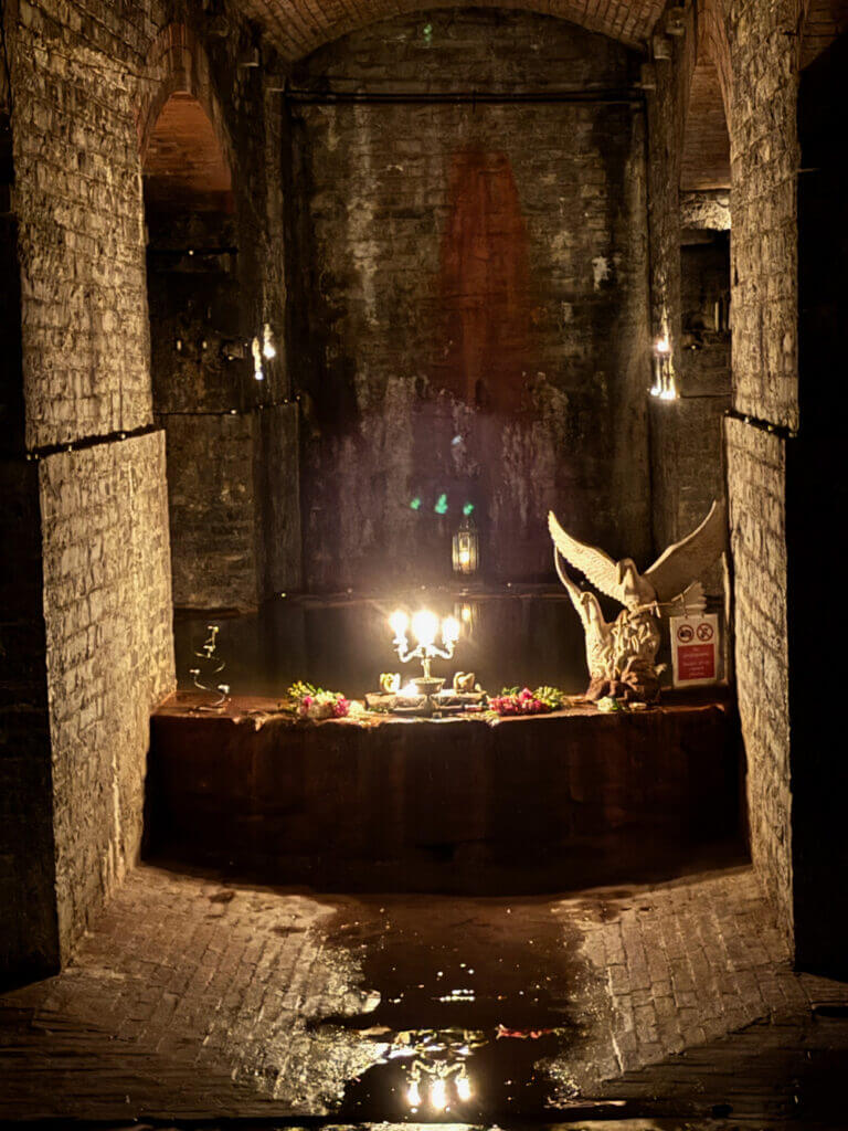 A dimly lit stone chamber with a central altar adorned with candles and a large sculpture, surrounded by reflective water puddles on the brick floor.