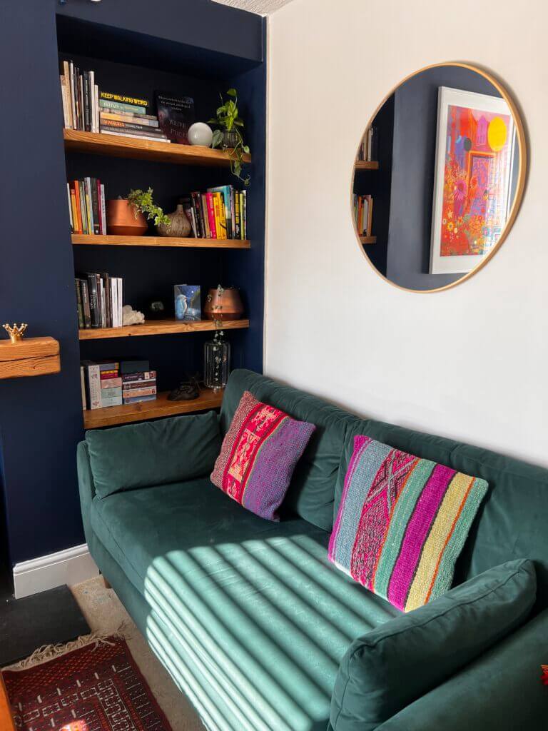 Cozy living room corner with a dark green sofa adorned with colorful pillows. A round mirror and a vibrant framed artwork are on the wall. Wooden shelves filled with books and plants are set into a dark alcove next to the sofa.