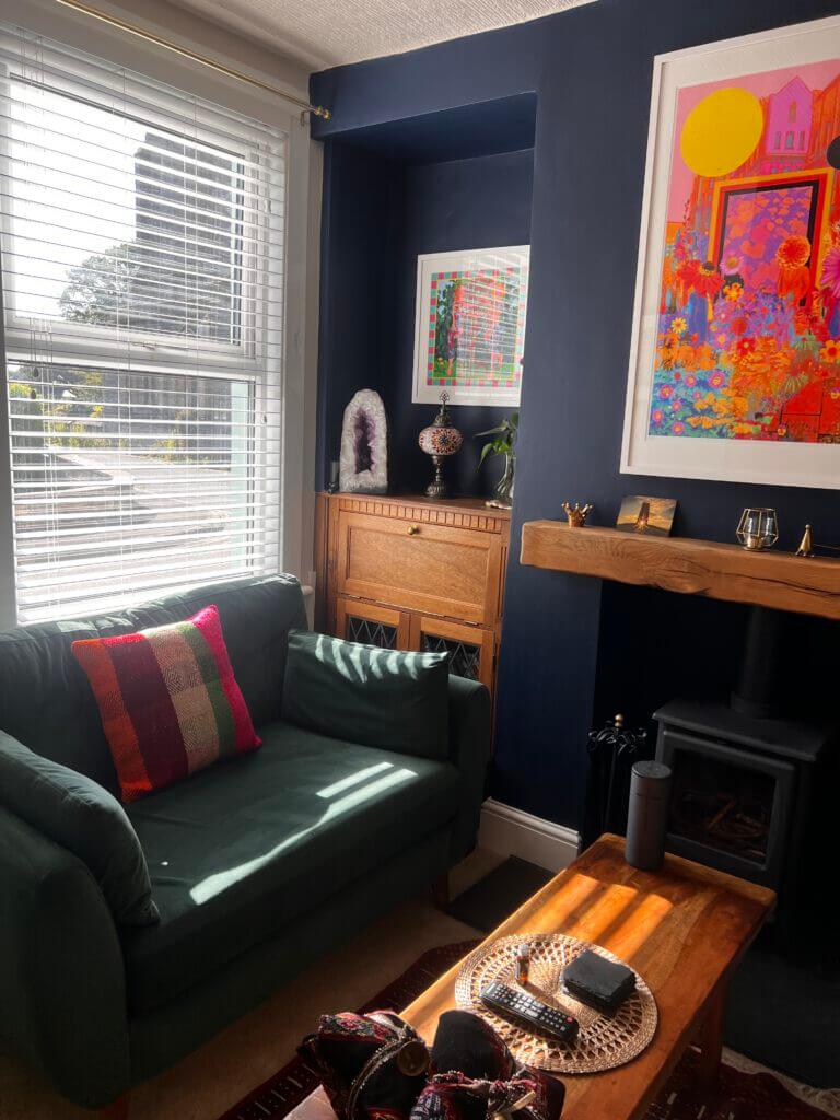 Cozy living room with a green sofa, colorful pillows, and a wooden coffee table. Sunlight filters through a window with blinds. A bright abstract painting hangs above a wooden mantle, with a small stove below. A cabinet and decorative items adorn the space.