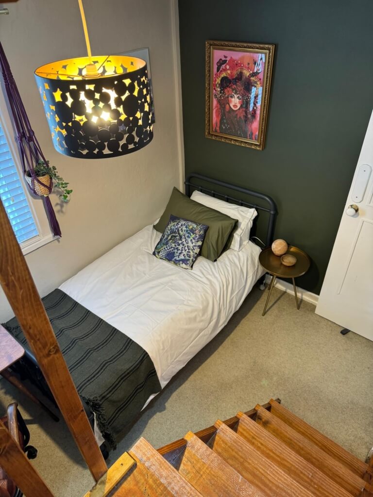 Cozy bedroom with a single bed, green and white bedding, a decorative pillow, and a small round side table. A star-patterned lampshade hangs from above, and a colorful artwork adorns the dark green wall. Stairs lead down to the carpeted floor.