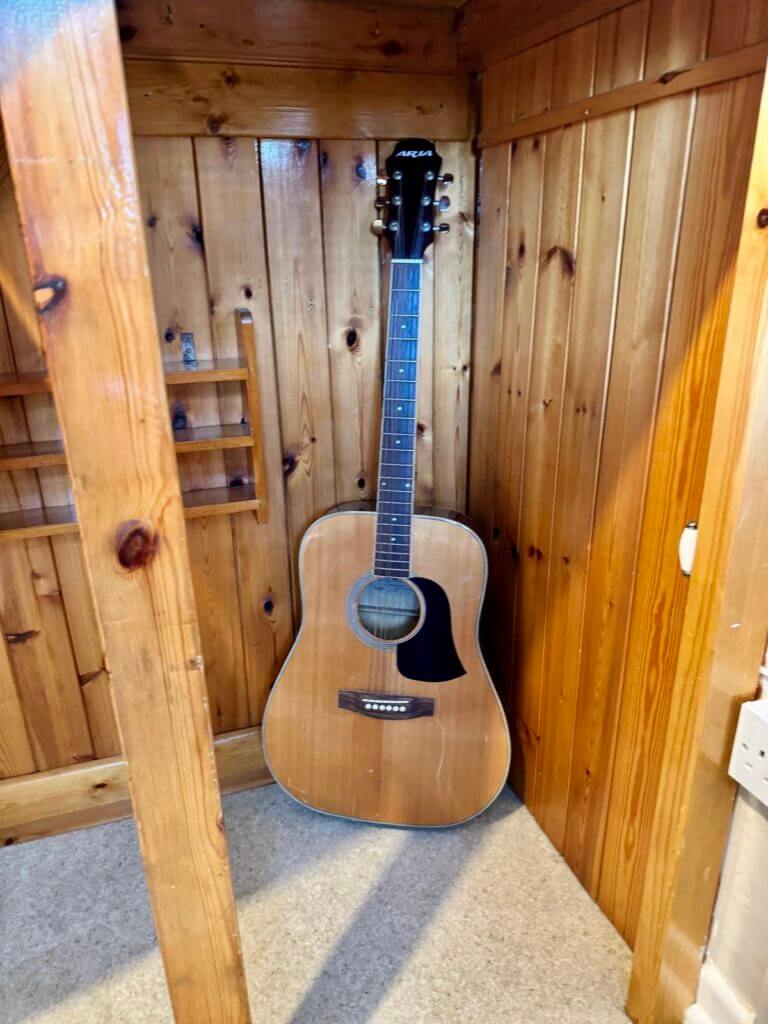 An acoustic guitar leaning against a wooden paneled wall in a corner of a room.