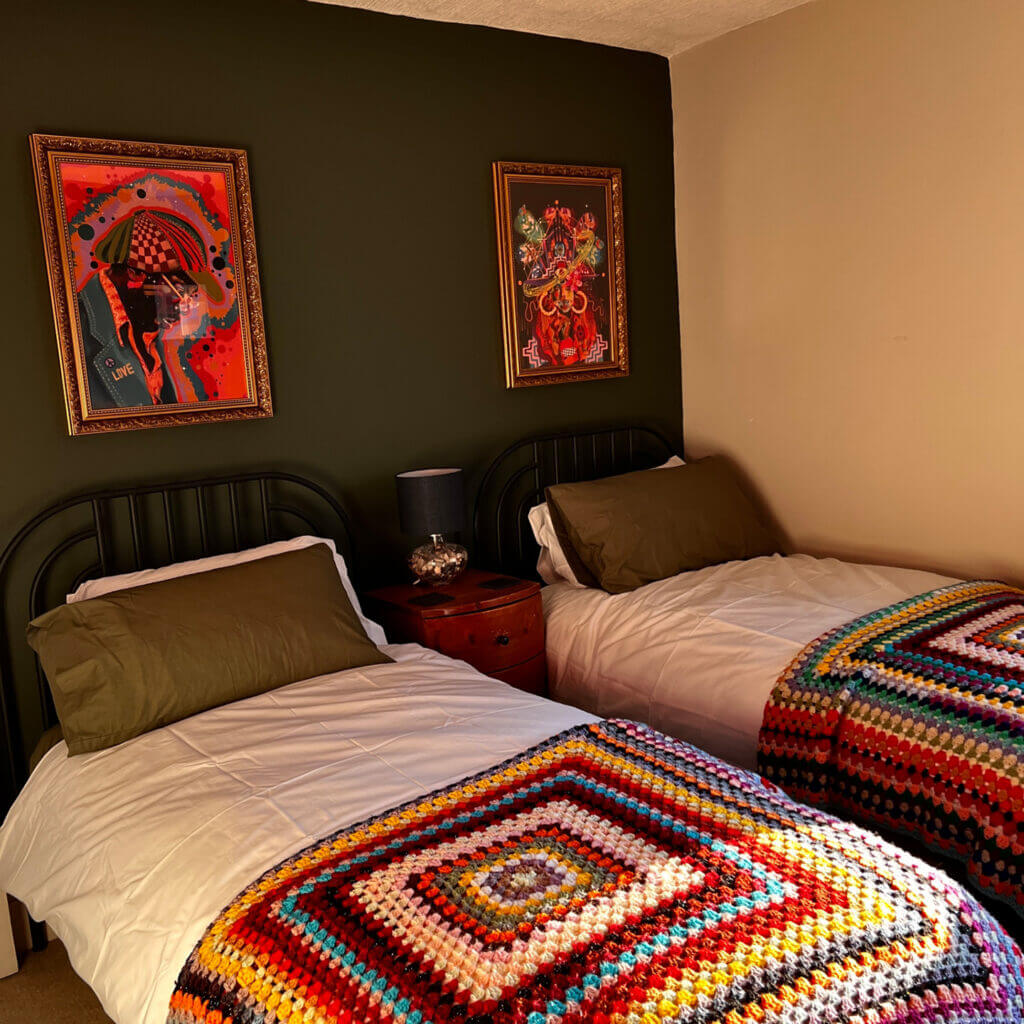 A cozy bedroom with two single beds, each adorned with colorful crochet blankets. A dark green accent wall features two vibrant framed artworks above a wooden nightstand holding a lamp.