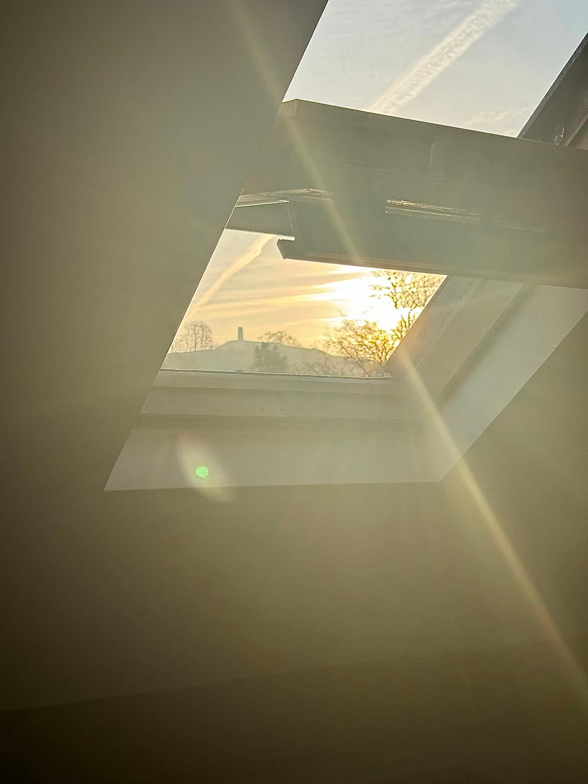 Sunlight streams through an open skylight, illuminating the room with warm tones. Outside, the sky is streaked with contrails, and the sun is low on the horizon, suggesting early morning or late afternoon.