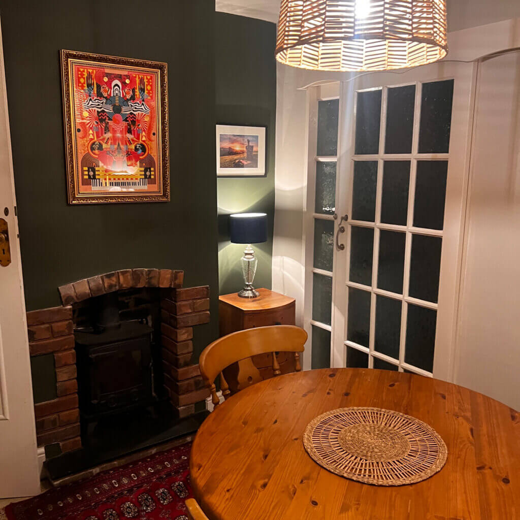 Cozy dining nook with a round wooden table, wicker lampshade, and small fireplace. Walls are decorated with artwork and a lamp sits on a side table near glass-paneled doors.