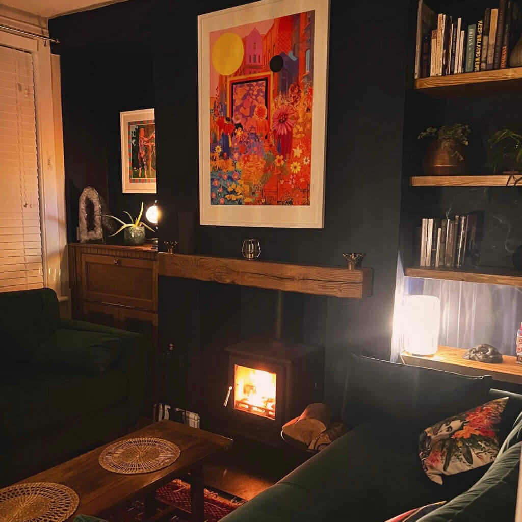 Cozy living room with dark walls, colorful artwork, and a small wood-burning stove. The room features green sofas, wooden shelves with books, and soft lighting, creating a warm, inviting atmosphere.