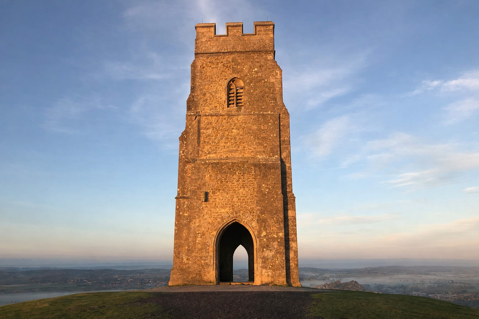 Stone tower on a grassy hilltop under a blue sky, with an arched doorway and battlements.