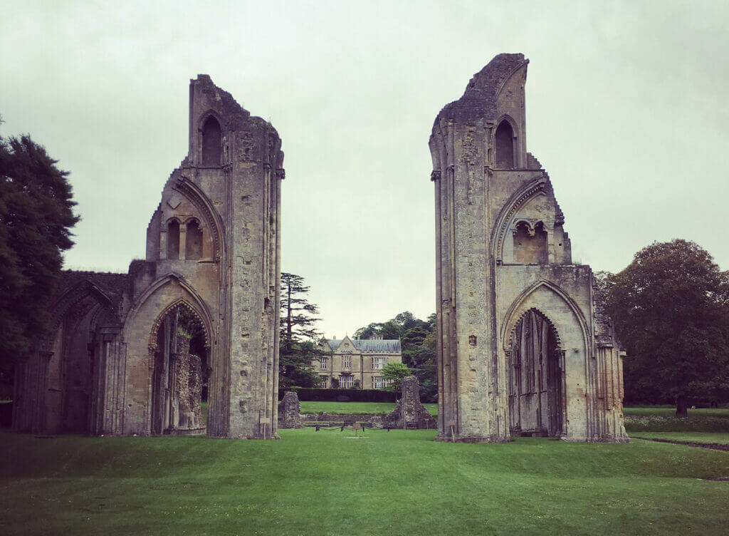 Ancient stone arches of Glastonbury Abbey stand amid green grass, part of a ruined structure. A large building is visible in the background, framed by the arches.