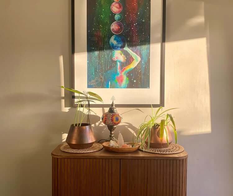 A wooden cabinet with potted plants and decor sits beneath a colorful framed artwork depicting a meditating figure and planets, bathed in warm sunlight.